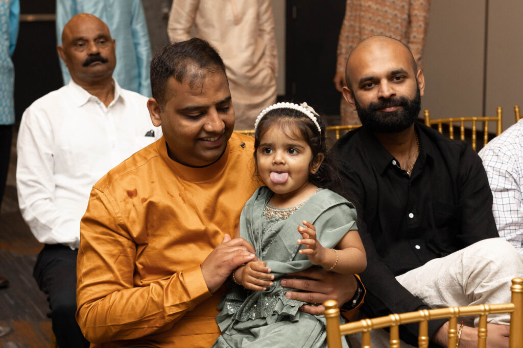 Father and his daughter sitting during a ceremony. Daughter has tongue sticking out and father is looking at her.