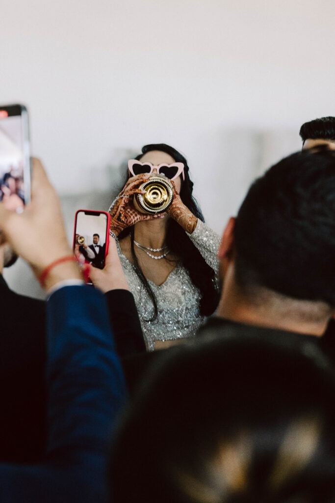 a bride in traditional indian attire drinking champagne out of the bottle while her friends take photos on their phones.