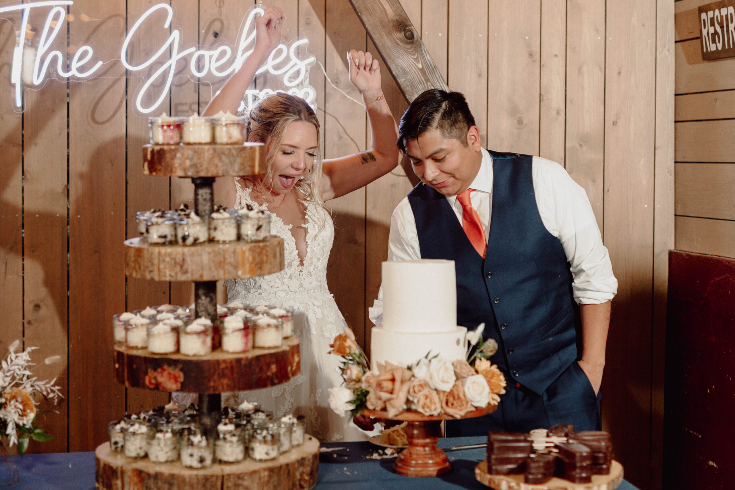 Bride and Groom at their dessert table. They are cutting the cake. Bride has her hands in the air, and their neon sign is behind them