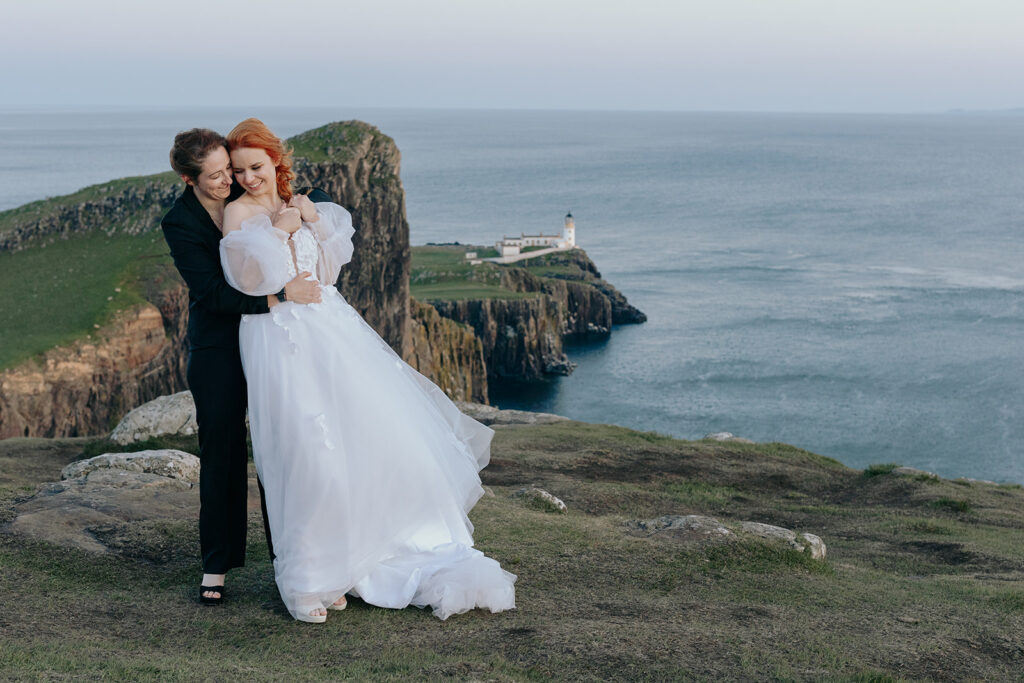 two brides at neist point in scottland uk. The bride wearing a wedding dress is leaning into the bride wearing a black suit.