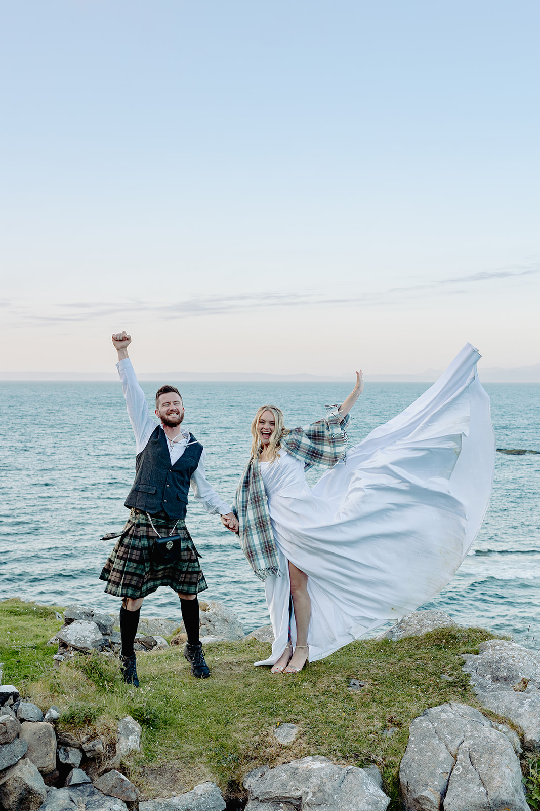 bride and groom with sea water behind them. They are wearing traditional Scottish attire and looking at the camera. The groom has his fist in the air and the bride is throwing her dress' train in the air.
